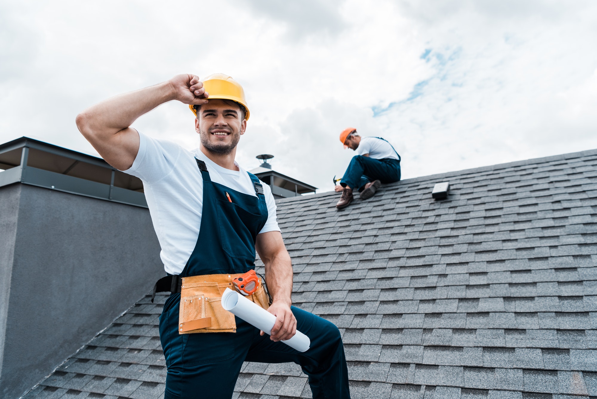 selective focus of happy repairman holding rolled paper while coworker repairing roof